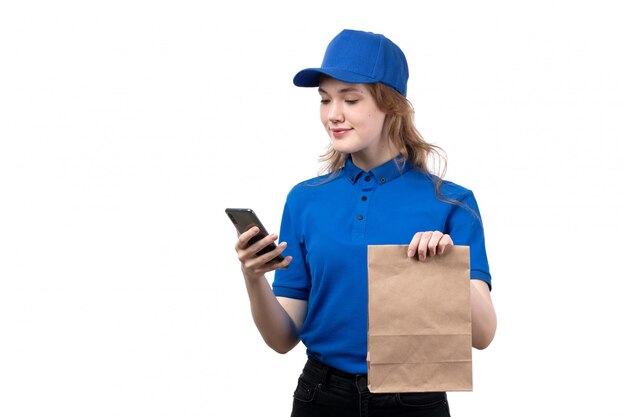 A front view young female courier female worker of food delivery service holding food package and using a phone with smile on white