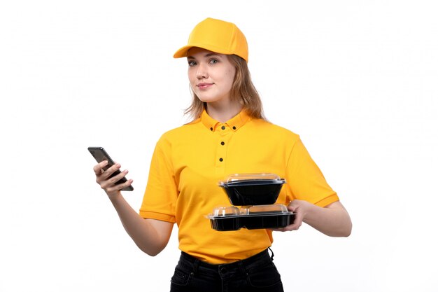 A front view young female courier female worker of food delivery service smiling holding food bowls and using a phone on white