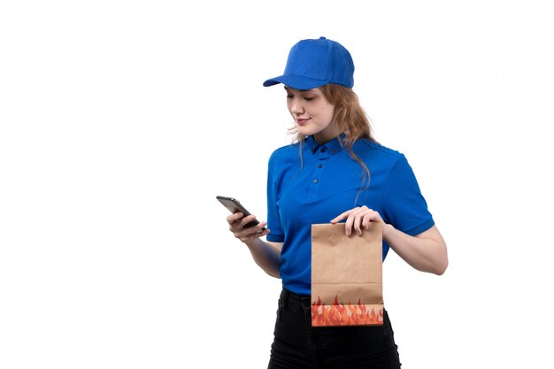 A front view young female courier female worker of food delivery service smiling holding package with food and using a phone on white