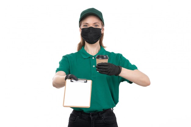 A front view young female courier in green uniform black gloves and black mask holding coffee cup and notepad