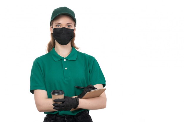 A front view young female courier in green uniform black gloves and black mask holding coffee cup and notepad