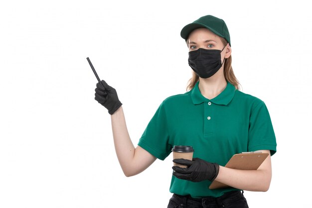 A front view young female courier in green uniform black gloves and black mask holding coffee cup and notepad