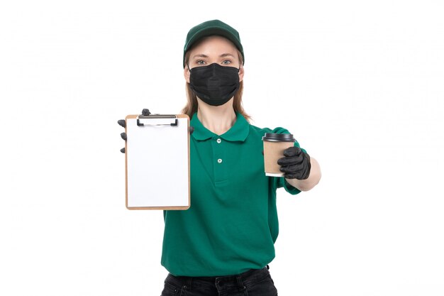 A front view young female courier in green uniform black gloves and black mask holding coffee cup and notepad