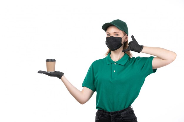 A front view young female courier in green uniform black gloves and black mask holding coffee cup