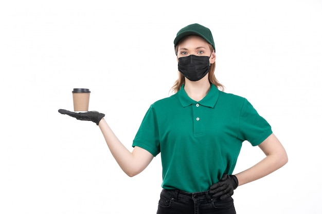 A front view young female courier in green uniform black gloves and black mask holding coffee cup