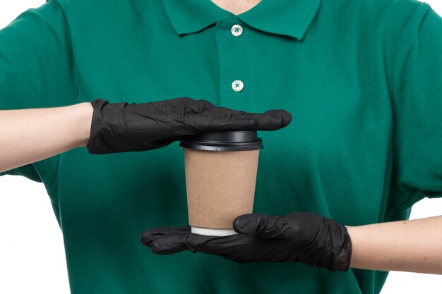 A front view young female courier in green uniform black gloves and black mask holding coffee cup