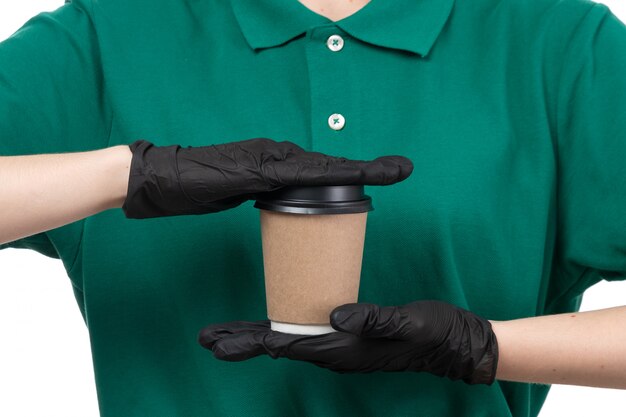 Молодая женщина-курьер в зеленой униформе, черные перчатки и черная маска, держащая чашку кофе, вид спереди
