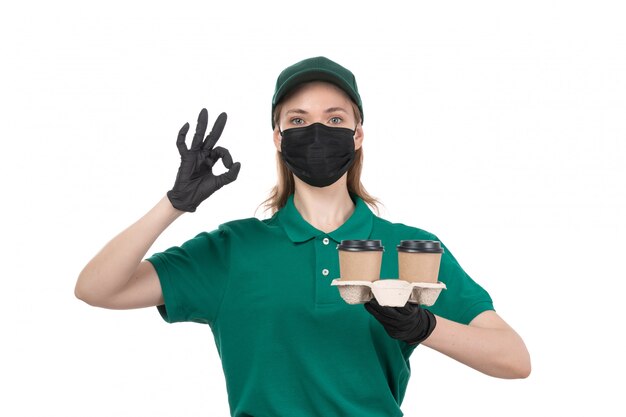 A front view young female courier in green uniform black gloves and black mask holding coffee cups