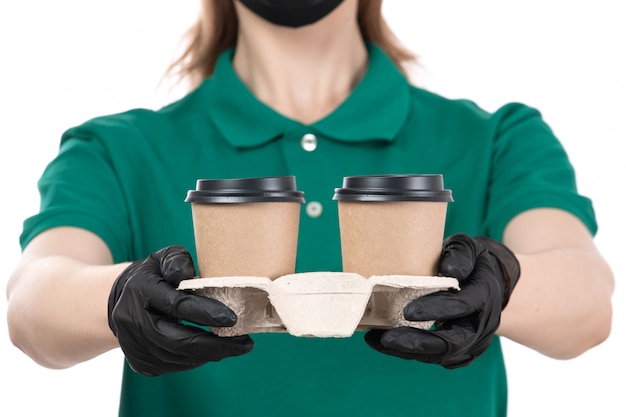 A front view young female courier in green uniform black gloves and black mask holding coffee cups