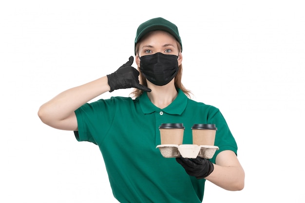 A front view young female courier in green uniform black gloves and black mask holding coffee cups