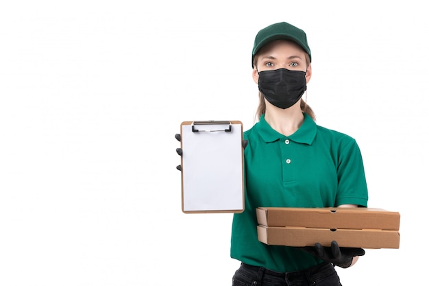 A front view young female courier in green uniform black gloves and black mask holding food delivery boxes