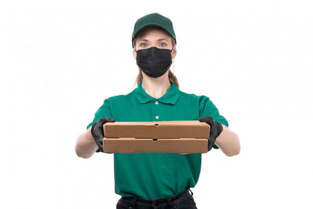 A front view young female courier in green uniform black gloves and black mask holding food delivery boxes