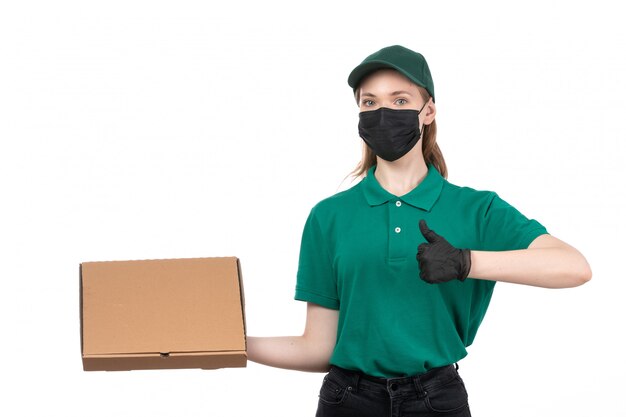 A front view young female courier in green uniform black gloves and black mask holding food delivery package delivering