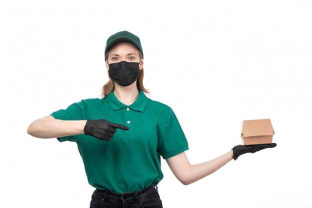 A front view young female courier in green uniform black gloves and black mask holding food delivery package delivering