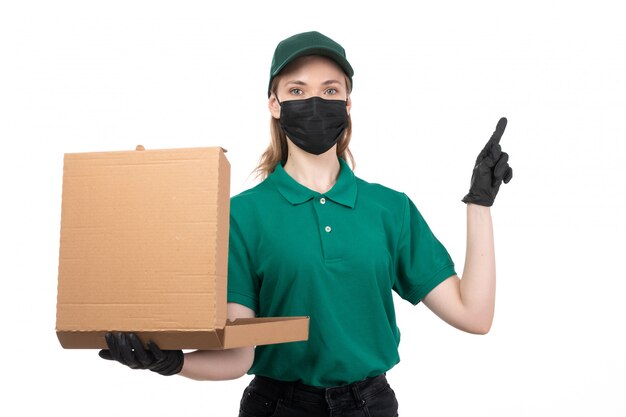 A front view young female courier in green uniform black gloves and black mask holding food delivery package delivering