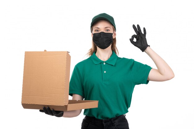 Вид спереди молодая женщина-курьер в зеленой форме, черные перчатки и черная маска, держащая доставку посылки с доставкой