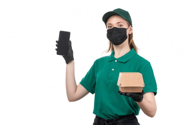 A front view young female courier in green uniform black gloves and black mask holding food delivery package and phone