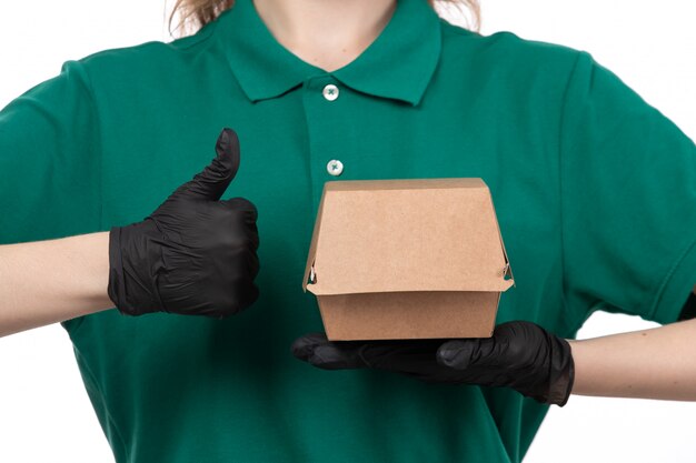 A front view young female courier in green uniform black gloves and black mask holding food delivery package