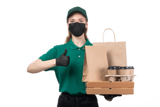 Вид спереди молодая женщина-курьер в зеленой форме, черные перчатки и черная маска, держащая доставку пакетов с доставкой еды