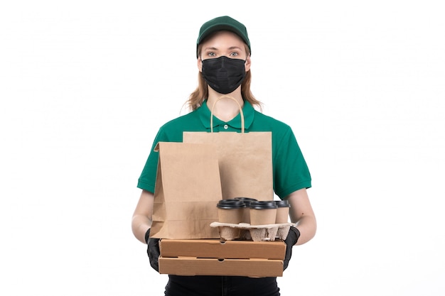 A front view young female courier in green uniform black gloves and black mask holding food delivery packages delivering