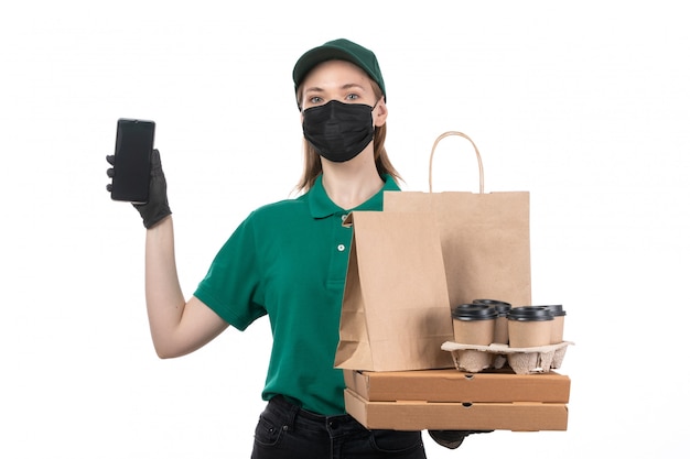 A front view young female courier in green uniform black gloves and black mask holding food delivery packages and phone delivering