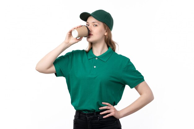 A front view young female courier in green uniform drinking coffee on white