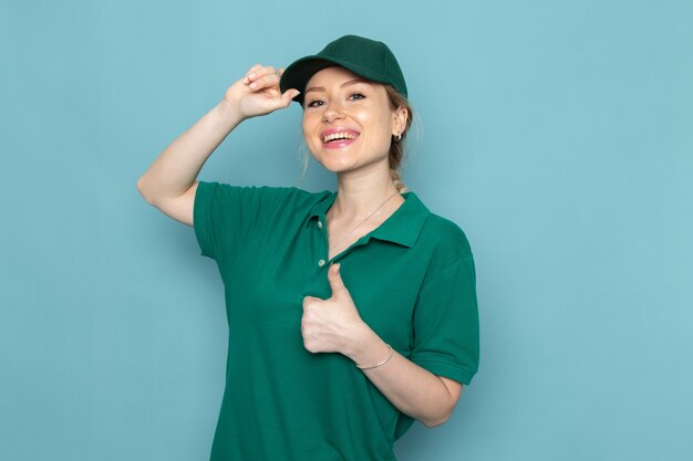 Front view young female courier in green uniform and green cape showing like sign with smile and posing on the blue space   job girl