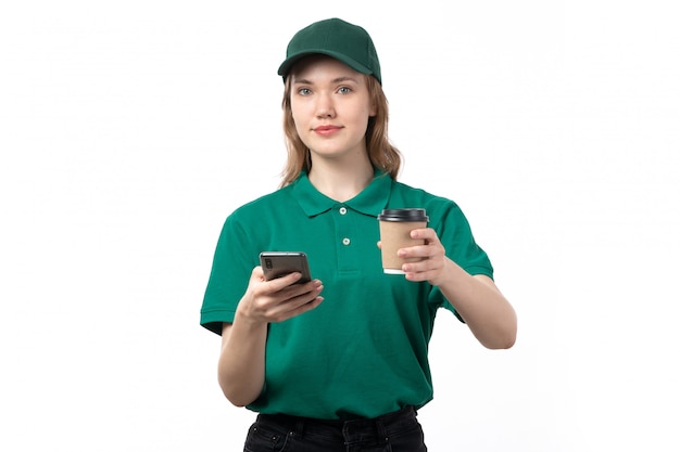 Вид спереди молодой женщины курьер в зеленой форме, держа чашку кофе и с помощью телефона на белом