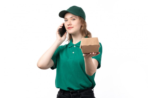 A front view young female courier in green uniform holding food package talking on the phone and smiling
