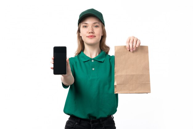 A front view young female courier in green uniform holding package with food and smartphone showing them