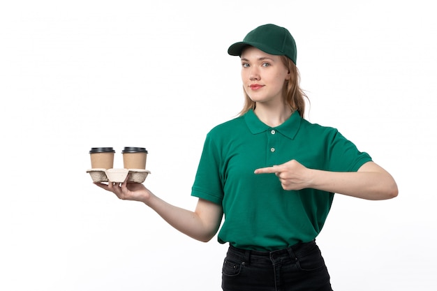 A front view young female courier in green uniform holding and pointing out coffee cups smiling on white