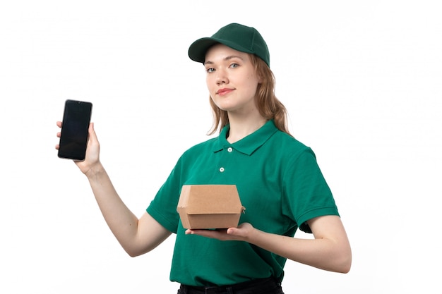 Молодая женщина-курьер в зеленой форме, держащая в руках смарптон и продуктовый пакет, вид спереди