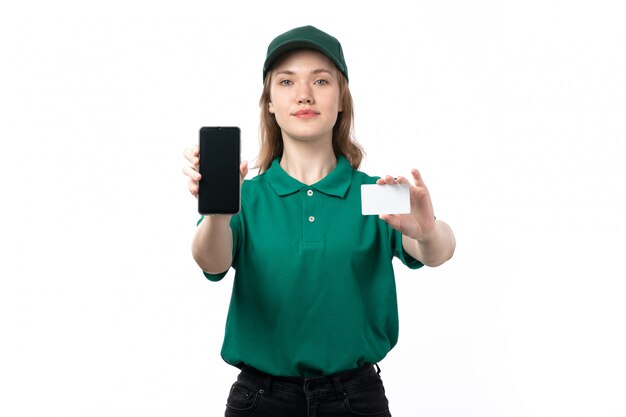 A front view young female courier in green uniform holding smartphone and white card smiling