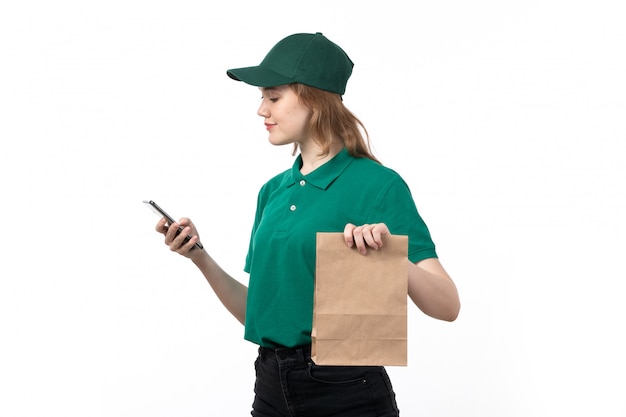 Молодая женщина-курьер в зеленой форме улыбается, держа смартфон, используя его вместе с пакетом продуктов