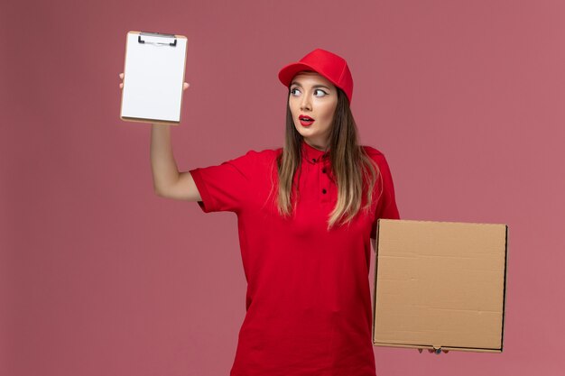 Бесплатное фото Вид спереди молодая женщина-курьер в красной форме, держащая коробку с едой для доставки с блокнотом на розовой форме службы доставки стола