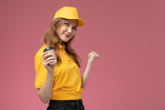Бесплатное фото Вид спереди молодая женщина-курьер в желтой форме, держащая чашку кофе на розовом фоне, служба доставки униформы
