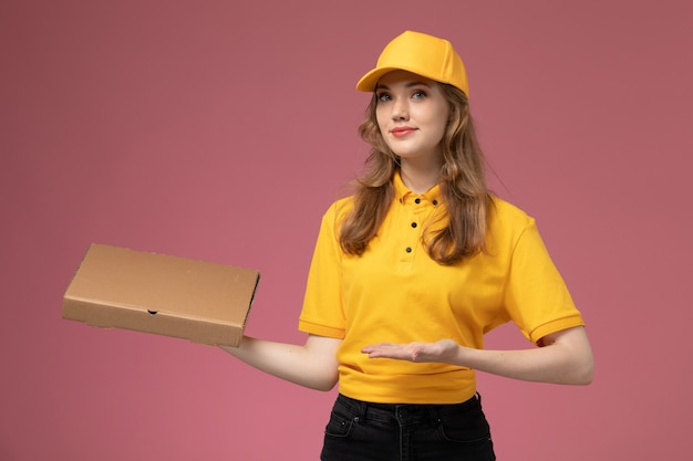 Бесплатное фото Вид спереди молодая женщина-курьер в желтой форме, держащая коробку с едой для доставки, улыбаясь на розовом столе, работник службы доставки униформы