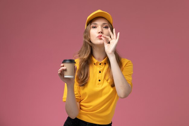 Бесплатное фото Вид спереди молодая женщина-курьер в желтой униформе с желтым плащом держит пластиковую кофейную чашку на темно-розовом фоне.