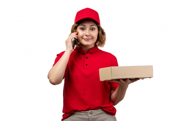 A front view young female courier in red shirt red cap holding delivery box smiling talking on the phone