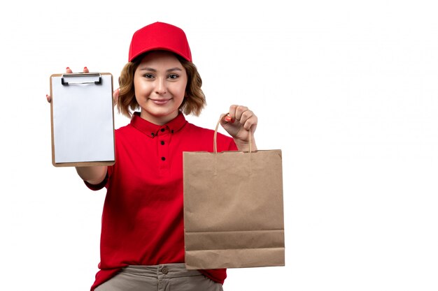 A front view young female courier in red shirt red cap holding delivery package and notepad smiling