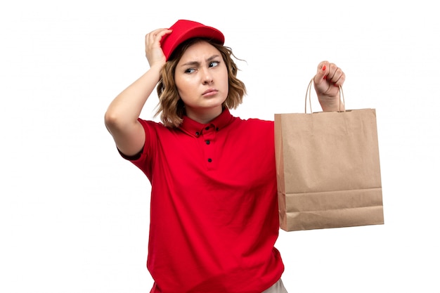 白い背景の均一なサービスを提供する上で配信パッケージを保持している赤いシャツの赤い帽子の正面の若い女性宅配便