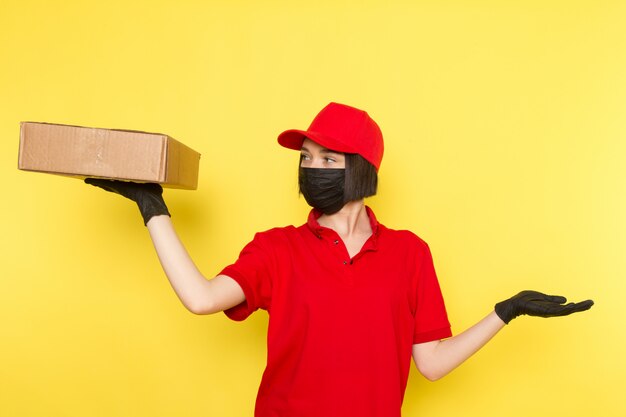 A front view young female courier in red uniform black gloves black mask and red cap holding food box