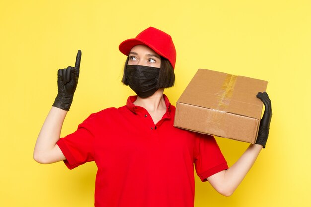 A front view young female courier in red uniform black gloves mask and red cap holding food box