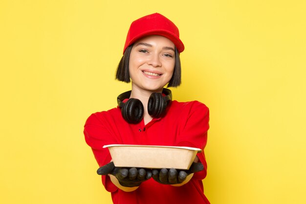 A front view young female courier in red uniform black gloves and red cap holding food bowl with smile