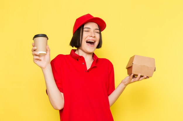 A front view young female courier in red uniform black gloves and red cap holding food box and coffee cup