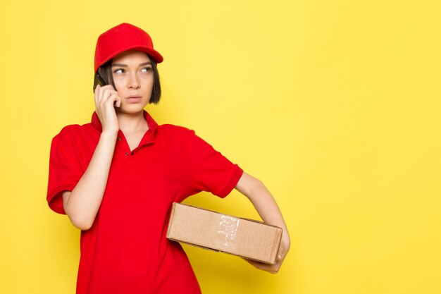 A front view young female courier in red uniform black gloves and red cap holding food box and talking on the phone