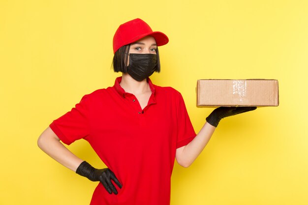 A front view young female courier in red uniform black gloves and red cap holding food box