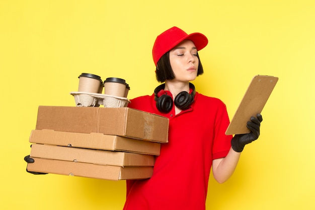 A front view young female courier in red uniform black gloves and red cap holding food boxes and coffee cups