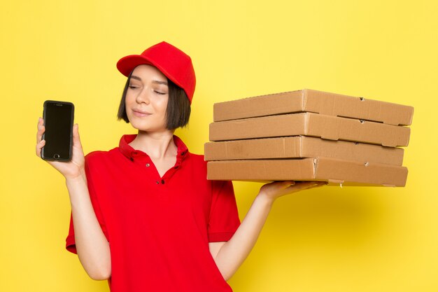 A front view young female courier in red uniform black gloves and red cap holding food boxes and phone