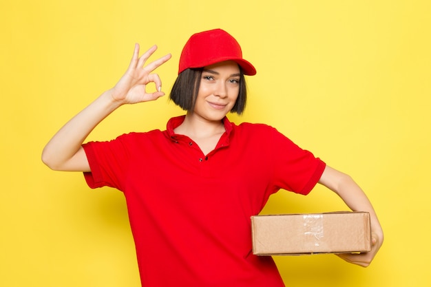 A front view young female courier in red uniform black gloves and red cap holding food package showing alright sign
